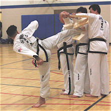 A black-belt kicking through a few boards, as three others hold the (now-broken) boards, looking away.